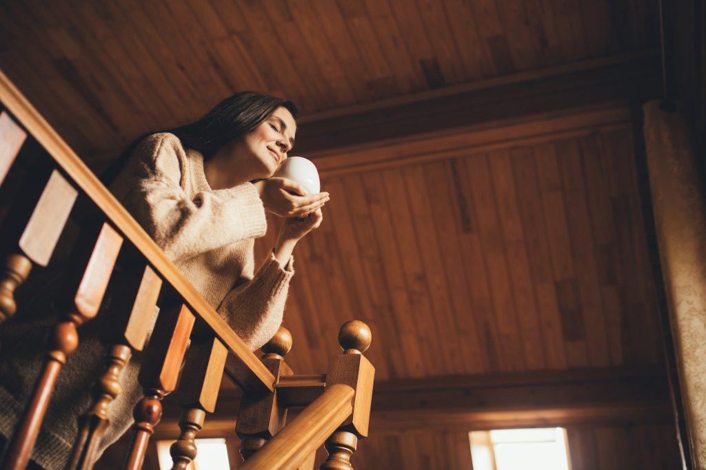 Young woman wearing warm sweater drinking hot tea in a cozy wooden country house.